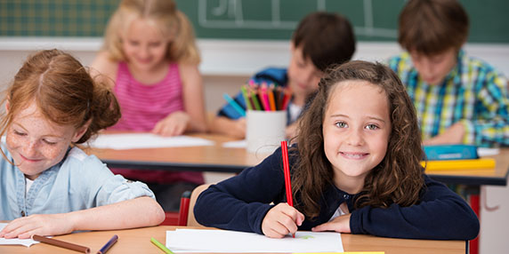 En flicka som sitter i ett klassrum tittar upp från sitt skolarbete. Hon ser glad ut.